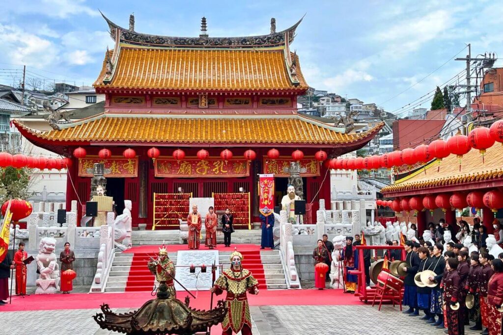 Nagasaki Confucius Temple Red and yellow Confucius Temple surrounded by red lanterns. Performers dressed in traditional attire are standing in front of the temple.