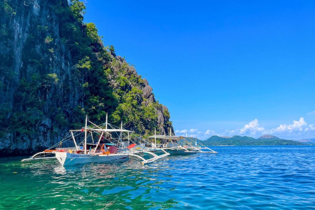 Coron Island Hopping Traditional Filipino boats anchored in front of limestone cliffs surrounded by blue-green water.