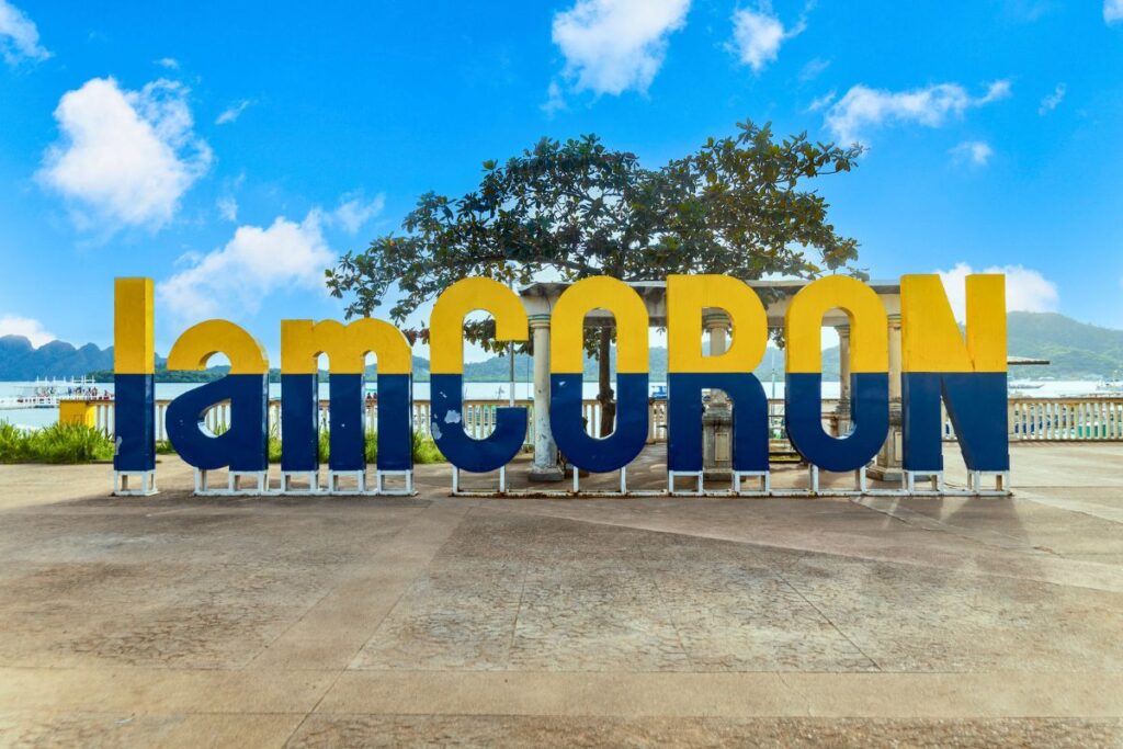 A blue and yellow sign that says "I am Coron" in front of the harbor in Coron Town.