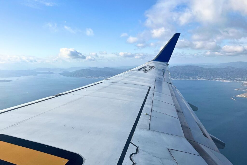 View from the window of a plane in flight. The plane's wing can be seen along with clouds and the ocean below.