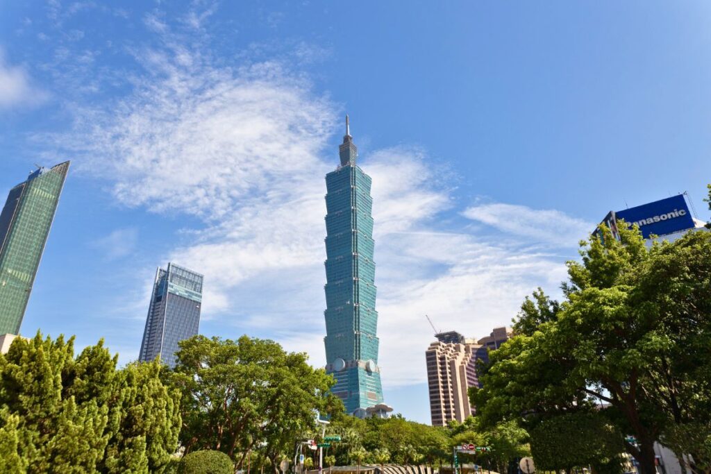 Taipei 101 building framed by green trees with blue partly cloudy skies in the background.