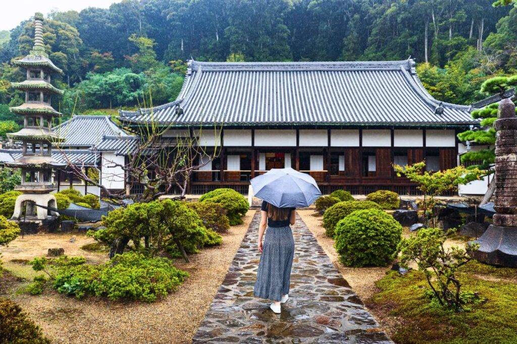 Krysti walking with an umbrella towards a Japanese temple surrounded by lush greenery.