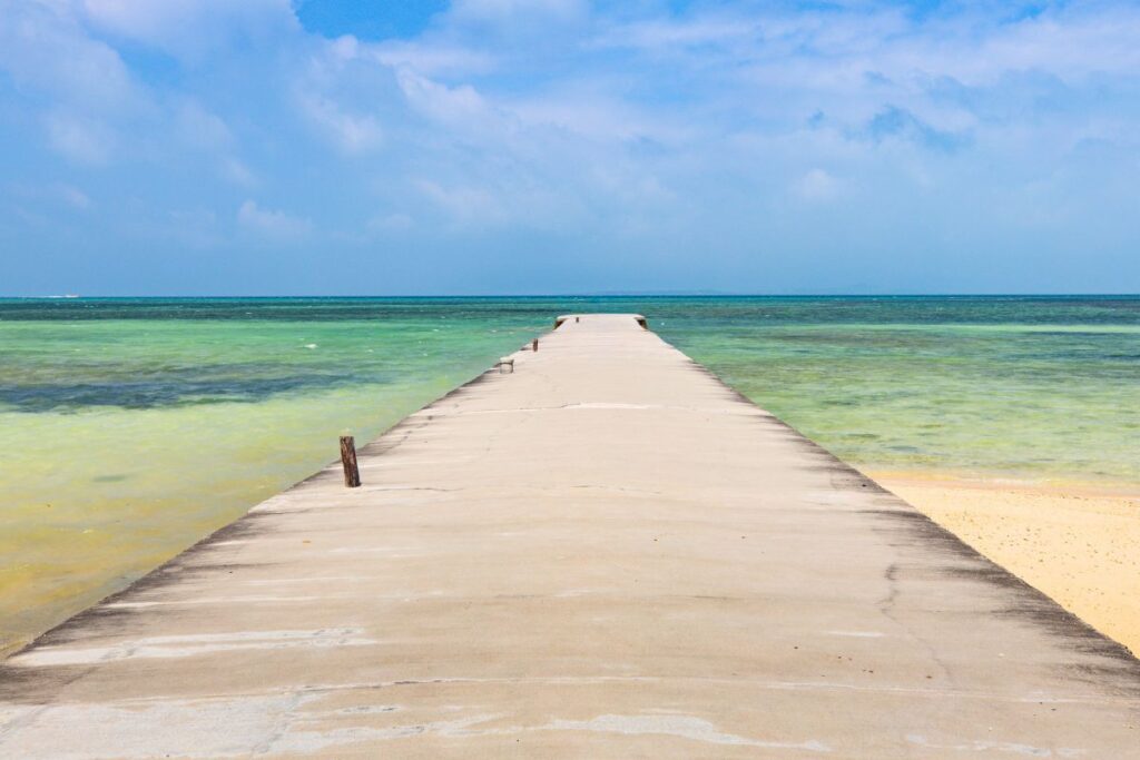 Concrete pier stretching out into the ocean surrounding by blue-green waters & sandy shores.