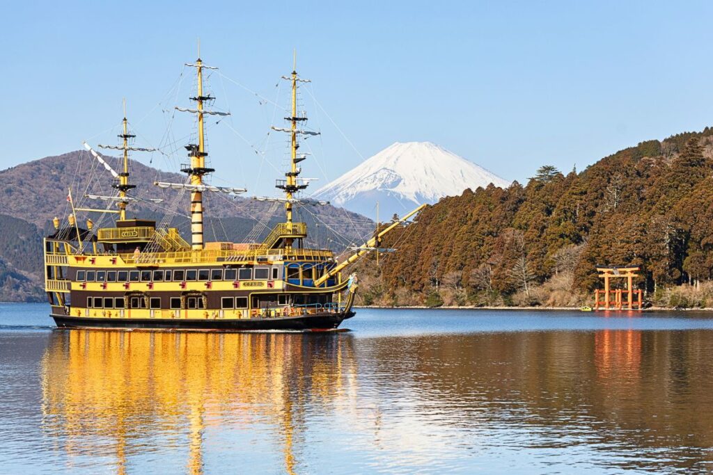 A view of Lake Ashi with Mt Fuji in the background, the Hakone Pirate Ship is sailing by, and the bright red Hakone torii gate sits along the shore.